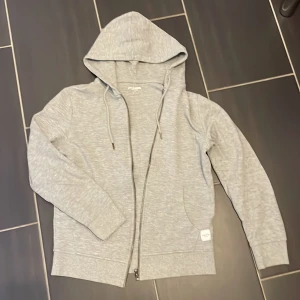 Grå hoodie från Jack & Jones - Snygg grå hoodie från Jack & Jones med dragkedja och huva. Tröjan har snörning vid halsen, två fickor framtill och ribbade muddar vid ärmslut och nederkant. Perfekt för chill dagar och enkel att matcha med jeans eller joggers.