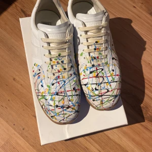 Maison Margiela sneakers med färgstänk - Unika vita sneakers från Maison Margiela med färgstänk i blått, rött, gult, grönt och svart. Skorna har klassisk låg siluett, snörning och gummisula. Tillverkade i skinn och har en konstnärlig vibe som sticker ut i mängden.