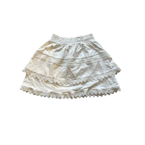 Vit kortkjol med spetsdetaljer - GinaTricot - Supersöt vit kortkjol med bred resår i midjan och flera lager med spetsdetaljer längs kanterna. Kjolen har en luftig och feminin look med volanger och passar perfekt till varma dagar. Materialet känns mjukt och bekvämt mot huden.