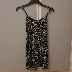 Svart blommig sommarklänning FB Sister - Supersöt svart kortklänning från FB Sister med vita blommor över hela tyget. Klänningen har smala axelband som går att justera, dragkedja i sidan och ärmlös design. Det finns snören man kan dra åt på baksidan (sista bilden) och en dragkedja man kan dra upp och ner när man ska ta på sig kläningen. Materialet är lätt och flowy, perfekt för varma dagar. Passformen är normal och modellen är feminin med lätt utställd kjol.