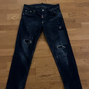 Mörkblå Dsquared2 jeans med slitningar - Äkta säljer ett par mörkblå jeans från Dsquared2 med coola slitningar och färgstänk på framsidan. Modellen har klassisk femficksdesign, gul Dsquared2-lapp bak och smal passform. Perfekt för dig som gillar streetstyle och detaljer. Pris kan diskuteras!