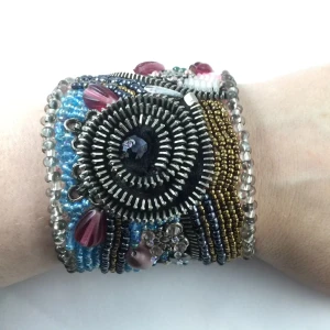 Färgstarkt pärlarmband med blommor - Unikt brett armband dekorerat med pärlor i blått, rosa, guld och silver. Armbandet har blommönster, glittriga detaljer och inslag av dragkedja som ger en cool kontrast. Perfekt statement piece för dig som gillar färg och kreativ design.