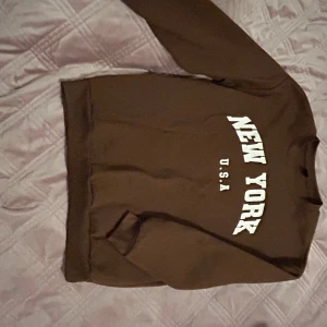 Brun sweatshirt New York från SHEIN - Brun sweatshirt från SHEIN med vit text 'NEW YORK U.S.A' på bröstet. Klassisk rund halsringning och långa ärmar. Mjuk insida och relaxed fit, perfekt för chill dagar. Tillverkad i polyesterblandning.