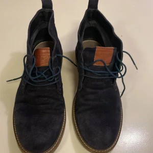 Mörkblå mockaboots från Gioseppo från Italien  - Snygga mörkblå boots från Gioseppo i mjuk mocka med klassisk snörning och bruna detaljer på plösen. Skorna har en platt sula i gummi och rund tå, perfekt för dig som gillar stilrena och bekväma skor med en modern touch.