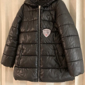 Svart pufferjacka från Benetton 10-12 år  - Svart pufferjacka från Benetton med huva och broderad Saint Moritz-patch på bröstet. Jackan är quiltad och har dragkedja framtill. Tillverkad i 100% polyester, perfekt för kalla dagar. Snygg och enkel design som håller dig varm.
