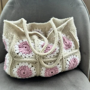Beige och rosa virkad axelväska - Superfin virkad axelväska i beige med virkade rutor i rosa och vitt. Väskan har rejäla handtag och ett unikt, retroinspirerat mönster som verkligen sticker ut. Perfekt för dig som gillar boho-stil och vill ha något personligt och mysigt.