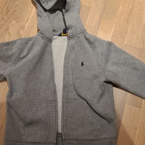 Grå hoodie från Polo Ralph Lauren - Klassisk grå hoodie från Polo Ralph Lauren med mörkblå logga broderad på bröstet. Tröjan har huva, dragkedja framtill och två fickor. Tillverkad i mjuk bomullsmix, perfekt för chill dagar. Enkel och stilren design som funkar året runt.Hoodien har två små prickar på armen därför priset.