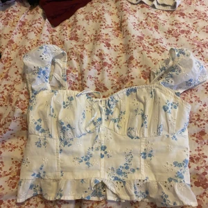 Blommig topp med volang från Hollister - Supersöt topp från Hollister i vitt med blått blommönster, volangkant nertill och broderade detaljer. Modellen är croppad med korta ärmar och knytning framtill. Perfekt för sommarens alla tillfällen! Storlek S och 100% bomull 
