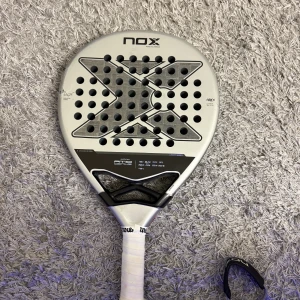 Nox AT10 Luxury Genius 18K Agustin Tapia - Nox AT10 Luxury Genius 18K padelracket signerat av Agustin Tapia. Tillverkad i 18K kolfiber för maximal prestanda. Racketen har sprickor på båda sidor av ramen, men greppet är i gott skick. Perfekt för dig som vill testa en toppmodell till ett lägre pris.