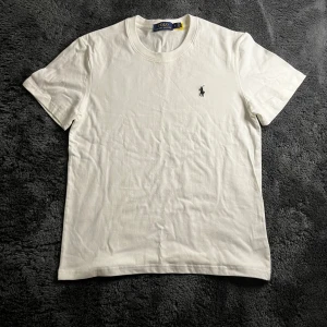 Vit t-shirt från Polo Ralph Lauren - Vit t-shirt från Polo Ralph Lauren med klassisk passform och rund hals. Ikoniska broderade loggan i svart på bröstet. Tillverkad i mjuk bomull som känns skön mot huden. Perfekt basic-plagg med stilren look.