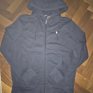 Polo Ralph Lauren hoodie zip - Snygg marinblå hoodie från Polo Ralph Lauren med dragkedja och huva. Tröjan har snörning vid huvan, klassisk logga på bröstet och praktiska fickor framtill. Perfekt för chill dagar och enkel att matcha med jeans eller joggers.