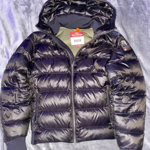Svart dunjacka Parajumpers Sheen - Snygg svart dunjacka från Parajumpers, modell Sheen. Jackan har glansig finish, huva, ribbade muddar och dragkedja framtill. Klassisk patch på ärmen och quiltad design som ger en cool look. Perfekt för kalla dagar när du vill hålla stilen.