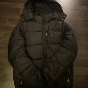 SÖKER BUD  - Svart pufferjacka från Polo Ralph Lauren med röd broderad logga på bröstet. Jackan har huva, dragkedja framtill och två sidofickor med dragkedja. Perfekt för kalla dagar och riktigt snygg streetstil. Fick i födelsedags present för nått år sen och har legat i garderoben, har ingen om äkta eller ej. 