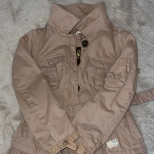 Beige jacka med bälte från Odd Molly - Snygg beige jacka från Odd Molly med markerad krage, stora knappar och bälte i midjan. Jackan har volangdetaljer nertill, fickor med lock och ett färgglatt paisleymönstrat foder. Perfekt för dig som vill ha en unik och cool look.