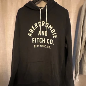 Svart hoodie Abercrombie & Fitch L - Svart hoodie från Abercrombie & Fitch i storlek L med vit text på bröstet och klassisk snörning vid huvan. Tröjan har lång ärm och en stor magficka framtill. Perfekt för chill dagar och enkel att matcha med jeans eller joggers.