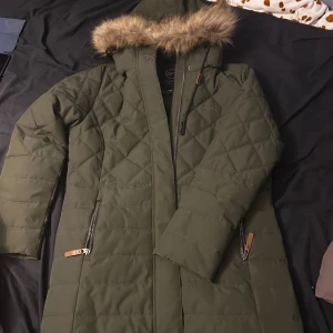 Mörkgrön quiltad dunjacka Five Seasons - Snygg mörkgrön lång dunjacka från Five Seasons med quiltat mönster, huva med fuskpäls och dragkedjor med läderdetaljer. Jackan har broderad logga på bröstet och metallmärke på ärmen. Perfekt för kalla dagar med sin varma och stilrena look.