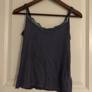 Lila linnetopp med spets från 157 - Säljer en lila linnetopp från The Black Label Collection, 157. Toppen har tunna axelband och en snygg spetskant längs ringningen. Perfekt att styla med jeans eller kjol för en chill look. Säljer då den inte används