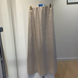 Beige satinkjol | Second Female - Fin långkjol i beige satin från Second Female. Kjolen har en rak och flowy passform med resår i midjan och dragkedja där bak. Observera bild 5!: kjolen har en tråd som sticker upp på framsidan. 