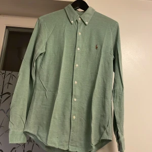 Grön skjorta från Polo Ralph Lauren - Snygg grön skjorta från Polo Ralph Lauren med klassisk button down-krage och broderad logga på bröstet. Skjortan är i bomull, har långa ärmar och vita knappar framtill. Perfekt för dig som gillar stilrena och tidlösa plagg. Obs .skjortan har ett litet hål på vänster axel och längre till höger som inte syns alls