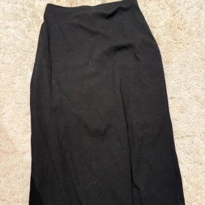 Svart långkjol i ribbad trikå - En svart långkjol i mjuk ribbad trikå med rak passform. Kjolen är enkel och stilren utan synliga detaljer, perfekt att matcha med olika toppar. Midjan är elastisk för extra komfort.