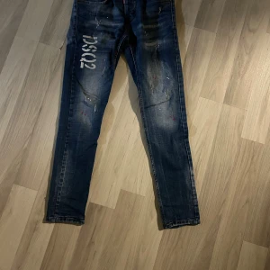 Dsquared2 blå skinny jeans med tryck - Snygga blå jeans från Dsquared2 med DSQ2-tryck på vänster lår, slitna detaljer och färgstänk. Modellen är skinny fit med klassiska fem fickor och dragkedjegylf. Perfekta för dig som gillar streetstyle och vill sticka ut.