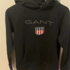 Svart hoodie från GANT med logga - Svart hoodie från GANT med broderad logga och amerikanskinspirerat emblem på bröstet. Klassisk känguruficka framtill och huva. Perfekt för en avslappnad och sportig stil.