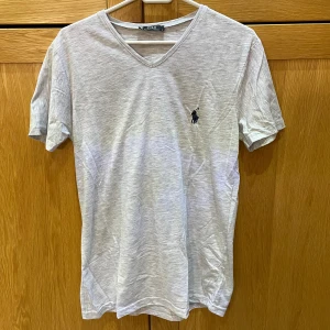 Ljusgrå t-shirt från Polo Ralph Lauren - Snygg ljusgrå t-shirt från Polo Ralph Lauren med v-ringning och klassisk broderad logga på bröstet. Tillverkad i mjuk bomull och har en normal passform. Perfekt för en clean och stilren look.