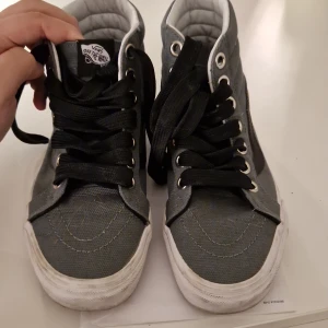 Grå höga Vans sneakers strl 39 - Säljer ett par klassiska Vans sneakers i grått tyg med svarta detaljer och svarta skosnören. Modellen är hög och har den ikoniska vita sulan med gummisula undertill. Perfekta för dig som gillar streetstyle och skatekänsla.Den som köper betalar för frakt även om det ska till ett anat land 