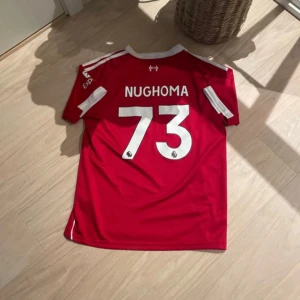 Liverpool FC röd fotbollströja Adidas - Riktigt snygg Liverpool FC matchtröja från Adidas i rött med vita detaljer och tryck. Kortärmad modell med klassiska Adidas-ränder på axlarna, klubbmärke och sponsortryck på bröstet. Materialet är lätt och andas, perfekt för träning eller match. Nummer 73 och namnet Nughoma på ryggen. Passar perfekt för 158 - 164