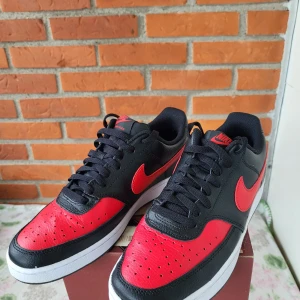 Nike Court Vision Low svart/röd - Nike Court Vision Low sneakers i svart och rött skinn med klassisk röd swoosh och svart snörning. Låg modell med vit platt sula och perforerad tå för extra ventilation. Perfekt för dig som gillar sportig och stilren look.