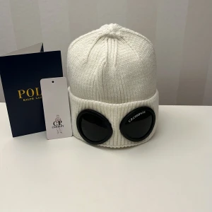 Vit C.P. Company mössa med goggles - Säljer en vit ribbstickad mössa från C.P. Company med ikoniska svarta goggles framtill. Mössan är tillverkad i 100% bomull och har en uppvikt kant. Perfekt för dig som vill sticka ut med en unik och trendig accessoar.