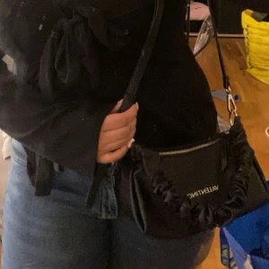 Svart handväska från Valentino - Snygg svart handväska från Valentino med guldig logga och dragkedja. Väskan har ett unikt rynkat handtag och är tillverkad i skinnimitation. Perfekt storlek för det viktigaste och har innerfack med dragkedja.OBS! Kan gå ner i pris