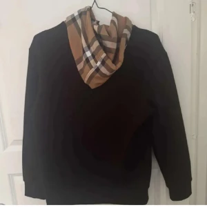 Svart hoodie med rutig huva - Svart hoodie med klassisk känguruficka och långärmad passform. Huvan har ett snyggt rutigt mönster i beige, brunt och svart, samt svarta snören. Perfekt för dig som vill ha en stilren men ändå unik look.