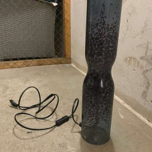  elektrisk lampa - Modern elektrisk lampa med svartmönstrad glasdesign och strömbrytare på sladden. Lampan är hög och cylinderformad, perfekt som dekorativ belysning. Mindre bruksspår men i gott skick. Glödlampa och exakt märke saknas.