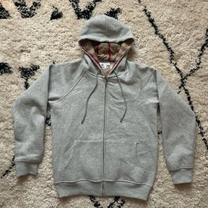Grå hoodie från Burberry med dragkedja - Säljer en grå hoodie från Burberry med klassiskt rutigt foder i huvan och snörning. Tröjan har dragkedja framtill, två fickor och ribbade muddar. Perfekt för dig som gillar stilren design med en touch av lyx. Är storlek M så den är för liten för mig.