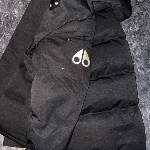 Svart dunjacka från Moose Knuckles - Svart dunjacka från Moose Knuckles med ikonisk metall-logga på ärmen, huva med pälsdetalj och flera fickor med dragkedja och knappar. Jackan har en puffad look och är perfekt för kalla dagar. Snygg och stilren design med detaljer i silver.