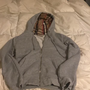 Grå hoodie med Burberry-mönstrad huva - Säljer en grå hoodie med dragkedja och snörning, där huvan har det klassiska Burberry-rutiga mönstret på insidan. Tröjan har två fickor framtill och är perfekt för dig som gillar streetstyle med en lyxig touch.