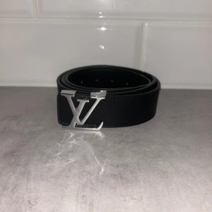 Svart Louis Vuitton skärp i skinn - Snyggt svart bälte Louis Vuitton med klassisk LV-logga i silver på spännet. Bältet är gjort i skinn och har diskret präglat monogram längs hela remmen. Perfekt accessoar för att lyfta din outfit med en lyxig touch.