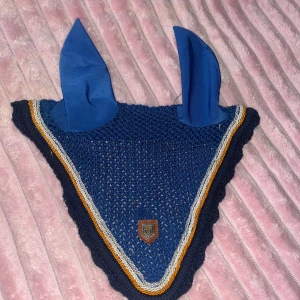 Blå huva från Trinity - Snygg blå ridhuva med långa tygöron och virkade detaljer. Huva med kantband i vitt, gult och blått samt ett litet märke framtill. Perfekt för häst och ridning, både snygg och praktisk. Materialet är stickat och tyg.