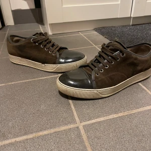 Bruna sneakers från Lanvin med svart tå - Snygga bruna sneakers från Lanvin med svart lackad rund tå och vit platt sula. Ovandelen är i mocka med snörning och metallöglor. Klassisk och stilren design som passar perfekt till jeans eller chinos. Märket syns tydligt under sulan.