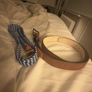 Brunt skärp i skinn 110cm & blått flätat bälte 110cm - Två snygga bälten säljes! Ett klassiskt brunt skärp i skinn med enkel design och ljus insida, perfekt till jeans eller chinos. Det andra är ett blått och vitt zigzag-mönstrat bälte med bruna detaljer och metallspänne, riktigt coolt och sticker ut i outfiten.