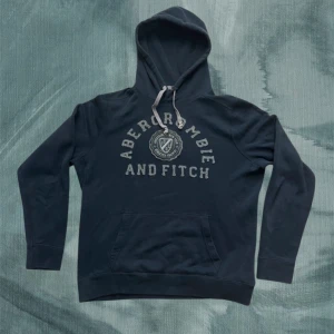 Mörkblå hoodie från Abercrombie & Fitch - Mörkblå hoodie från Abercrombie & Fitch med broderad logga och text framtill. Tröjan har huva med snörning, känguruficka och långa ärmar. Perfekt för en chill och avslappnad stil nu till hösten/vintern. Pris kan diskuteras vid snabb affär! Ha det gött// Gidlöfs
