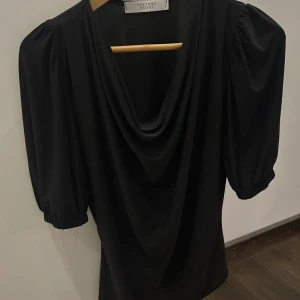 Svart kortärmad blus  - Svart blus från Sisters Point, köpt på salt, med snygg draperad urringning och puffiga halvlånga ärmar. Den har en stilren look och är tillverkad i ett mjukt, följsamt material som ger en skön passform.