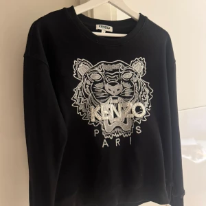 Svart Kenzo sweatshirt med tigerbrodyr - Svart sweatshirt från Kenzo med ikonisk silverbroderad tiger och logga på bröstet. Tröjan har rund hals, långa ärmar och en relaxed passform. Perfekt statement-plagg för dig som gillar streetstyle och snygga detaljer.