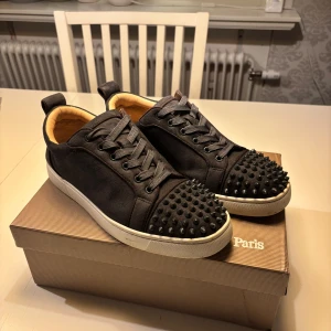 Christian Louboutin gråa & vita sneakers med nitar - Gråa/vita sneakers från Christian Louboutin i mocka med coola svarta nitar på tån och klassisk röd sula. Skorna har snörning, vit platt sula och beige innersida. Kommer med originalkartong och dustbag. Perfekt för dig som vill sticka ut med lyxig streetstyle.