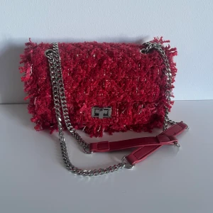 Röd axelväska med kedja - Snygg röd axelväska med fluffig, texturerad yta och silverfärgad kedja som axelrem. Väskan har ett coolt twistlås i metall och detaljer i rött läder på remmen. Perfekt för dig som vill sticka ut med en unik och trendig väska.