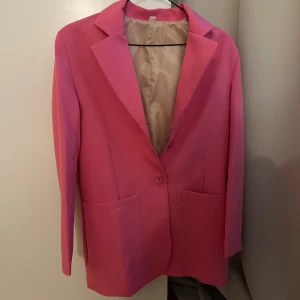 Rosa kavaj med enkel knapp - Snygg rosa kavaj med klassisk krage och en enkel knapp framtill. Kavajen har två stora fickor och är helfodrad med ljus insida. Perfekt för dig som vill sticka ut med färg och stil. Passar till både jeans och kostymbyxor.