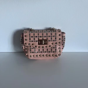 Rosa axelväska med nitar och kedja - Snygg rosa axelväska med fyrkantigt mönster och coola guldfärgade nitar. Väskan har en kedja som axelrem och ett metallspänne framtill. Materialet är syntet och detaljerna ger en edgy vibe. Perfekt för dig som gillar att sticka ut med accessoarer.