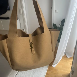 Beige axelväska  - Snygg och rymlig axelväska från i beige mocka med ikoniskt spänne i guld framtill. Väskan har breda handtag och stilren design som gör den enkel att matcha med olika outfits. Perfekt för dig som gillar lyxiga accessoarer med klassisk touch.