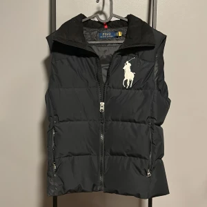 Svart dunväst från Polo Ralph Lauren - Säljer nu denna extremt sällsynta dunvästen från polo Ralph Lauren. Finns inga defekter eller liknande att den är använd. Ny skick 10/10.Följer med en luva. Qr kod fungerande. Pris går att diskutera. Tveka inte på att höra av dig.